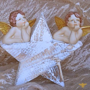 May include: A decorative star with a distressed white finish, featuring two cherub figurines with golden wings and reddish-brown hair. The cherubs are resting their chins on their hands, creating a serene and angelic scene.