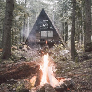 May include: A campfire burns brightly in front of a wooden A-frame cabin in the woods. The cabin has large windows and a wooden porch.
