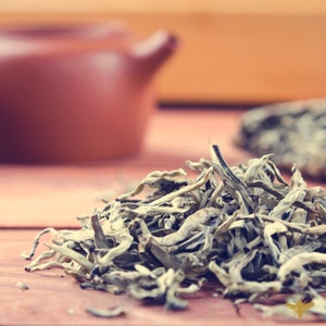 May include: A close-up of a pile of loose leaf white tea, with a brown teapot and a tea cake in the background.