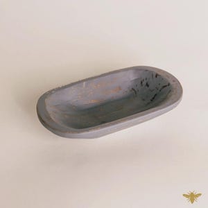 May include: A gray, oblong wooden bowl with a distressed finish. The bowl has a shallow, open design and is likely used for decorative purposes or serving small items. The wood grain is visible, adding to its rustic charm.