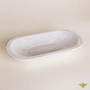 May include: A white, oval-shaped wooden bowl with a distressed finish. The bowl has a shallow interior and a slightly raised rim. The wood grain is visible through the white paint, giving it a rustic appearance. The bowl is empty and sits on a neutral background.