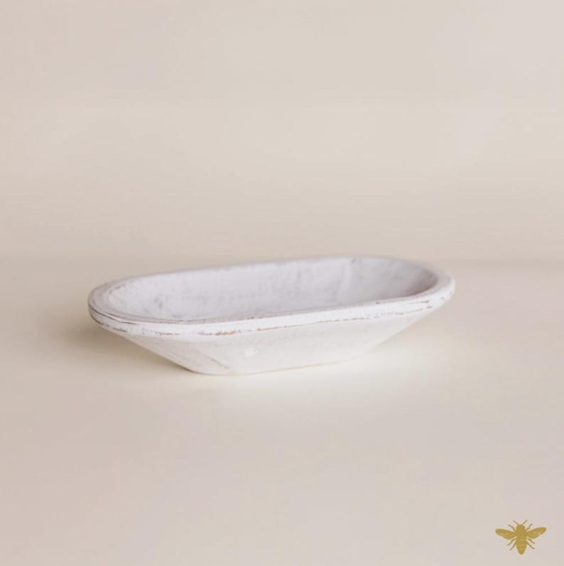 May include: A white, oval-shaped wooden bowl with a distressed finish. The bowl has a shallow depth and a slightly raised rim. The wood grain is visible through the white paint, giving it a rustic appearance. The bowl is empty and sits on a neutral surface.