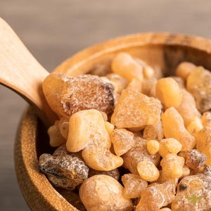 May include: Close-up of a wooden bowl filled with amber-colored frankincense resin pieces. A wooden spoon rests in the bowl, partially visible. The resin pieces vary in size and shape, with a mix of light and dark tones.
