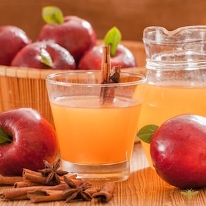 May include: A glass pitcher filled with apple cider, surrounded by red apples and cinnamon sticks. The cider is a light amber color and the apples are shiny and red. There are also cinnamon sticks and star anise on the table.