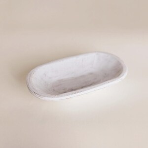 May include: A white, oval-shaped wooden bowl with a distressed finish. The bowl has a shallow depth and is ideal for decorative purposes or serving food. The wood grain is visible through the white paint, adding to its rustic charm.