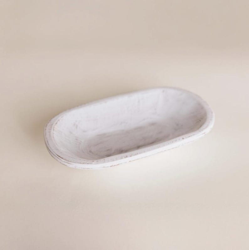 May include: A white, oval-shaped wooden bowl with a distressed finish. The bowl has a shallow depth and is ideal for decorative purposes or serving food. The wood grain is visible through the white paint, adding to its rustic charm.