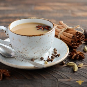 May include: A cup of chai tea with star anise floating on top, served in a speckled white mug and saucer. Cinnamon sticks, cloves, and cardamom pods are arranged around the cup on a wooden surface, creating a warm and inviting scene.