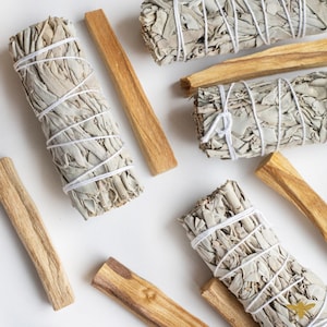 May include: Overhead shot of white sage bundles tied with white string and light brown palo santo sticks on a white surface. The sage bundles are a light green color. The palo santo sticks are a light brown color.