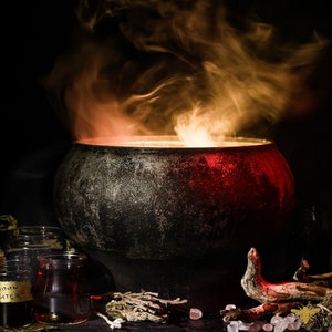 May include: A black cauldron with steam rising from it, surrounded by dried herbs, twigs, and small pink sweets. The cauldron is lit from within, casting a warm glow.