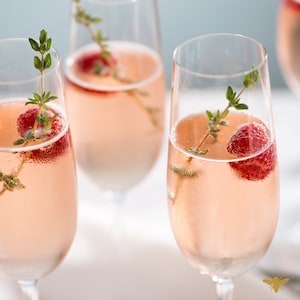 May include: Three champagne flutes filled with a pink sparkling beverage and garnished with fresh raspberries and sprigs of thyme.