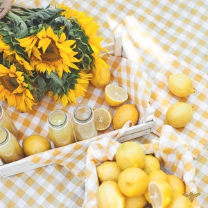 Puede incluir: Una caja de madera blanca llena de limones y limas, con cuatro botellas de vidrio de limonade sobre un mantel a cuadros amarillo y blanco. Un ramo de girasoles está en el fondo.
