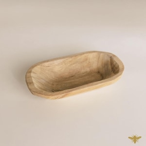 May include: A light-colored, wooden, oval-shaped bowl. The bowl has a smooth, natural finish and a shallow interior. The wood grain is visible, adding texture. The bowl is empty and sits on a neutral background.