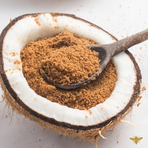 Puede incluir: Primer plano de una cáscara de coco llena de azúcar moreno. Una cuchara de madera está parcialmente sumergida en el azúcar.