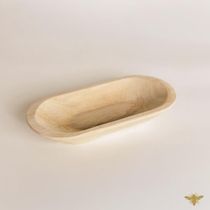 May include: A light-colored, oval-shaped wooden bowl. The bowl has a smooth, natural finish and a shallow interior. The design is simple and rustic, suitable for serving or display. The bowl is made of a light-colored wood.