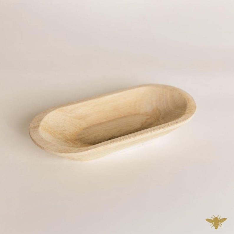 May include: A light-colored, oval-shaped wooden bowl. The bowl has a smooth, natural finish and a shallow interior. The design is simple and rustic, suitable for serving or display. The bowl is made of a light-colored wood.