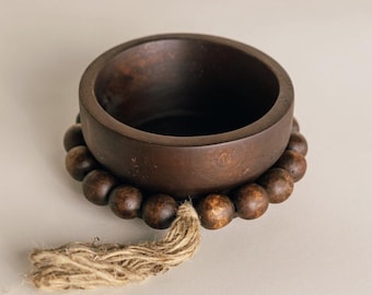 May include: A dark brown wooden bowl sits atop a beaded garland with a natural jute tassel. The bowl has a smooth, rounded shape and a dark interior. The beads are a similar color to the bowl.