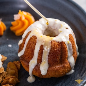 May include: A close-up of a small, round cake with a white glaze drizzled with a golden syrup. The cake is on a dark blue plate with a small orange swirl of frosting and a pile of brown, crispy crumbs.