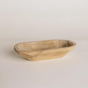 May include: A light-colored, wooden, oblong bowl with a shallow interior. The bowl has a smooth, natural finish and is likely hand-carved. The design is simple and rustic, suitable for decorative or functional use. The bowl is empty and sits on a neutral background.
