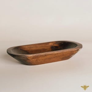 May include: A dark brown, oblong wooden bowl with a smooth, polished finish. The bowl has a shallow depth and gently curved sides, perfect for decorative or functional use. The wood grain is visible, adding to its rustic charm.