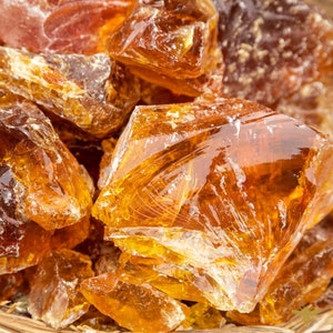May include: Close-up of a pile of amber-colored, translucent resin chunks. The pieces vary in size and shape, with sharp edges and a glossy surface. The light catches the facets, creating highlights and shadows.
