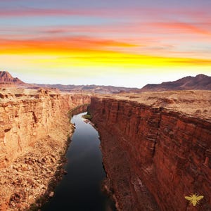 May include: A scenic landscape photograph of a deep canyon with red rock walls and a river flowing through it. The sky is a vibrant mix of yellow, orange, and pink hues, suggesting a sunrise or sunset. The image evokes a sense of vastness and natural beauty.