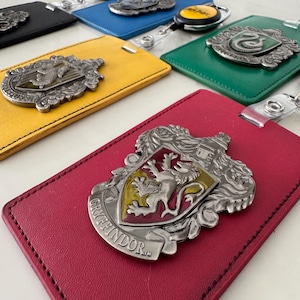May include: A collection of colorful luggage tags featuring various house crests. The tags are in red, yellow, green, blue, and black, each with a metal emblem and a clear plastic strap. The Gryffindor tag is in the foreground.
