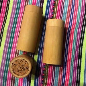 Peut inclure: Deux contenants en bambou naturel avec des couvercles en liège. Les contenants sont cylindriques et ont une finition lisse de couleur brun clair. Ils sont posés sur un tissu rayé coloré.