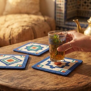 May include: Three square coasters featuring a geometric pattern in blue, cream, teal, and orange. Each coaster has a textured, woven appearance. A clear glass filled with tea and mint leaves sits on one of the coasters, held by a hand.