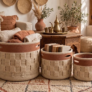 May include: A set of three round, woven storage baskets in varying sizes. Each basket features a cream-colored base with a textured, checkered pattern and a solid terracotta-colored rim with integrated handles for easy carrying.