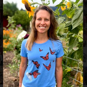 May include: A light blue t-shirt featuring a monarch butterfly design. The butterflies are orange and black, with some having white accents. The shirt is worn with white shorts. The background includes sunflowers and greenery.