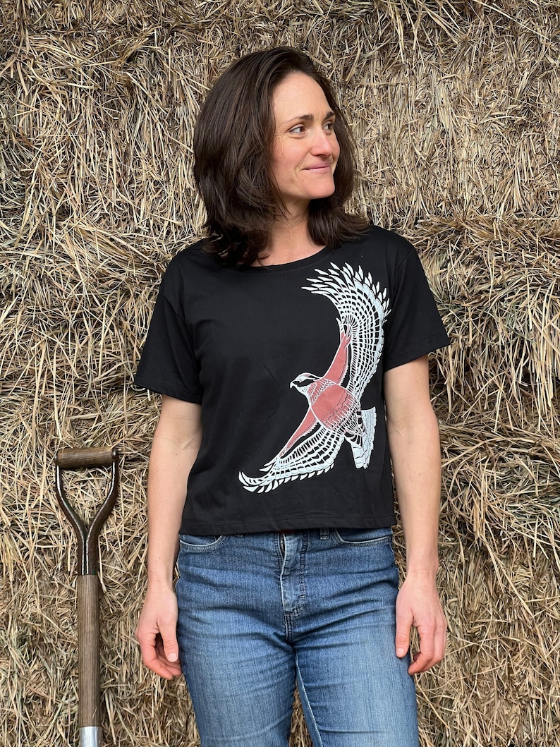 May include: A black cropped t-shirt with a white and red graphic of a hawk in flight. The shirt is worn by a person standing in front of a bale of hay.