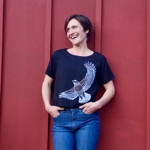 May include: A woman wearing a black t-shirt with a white bird graphic and blue jeans. She is standing in front of a red wooden wall.