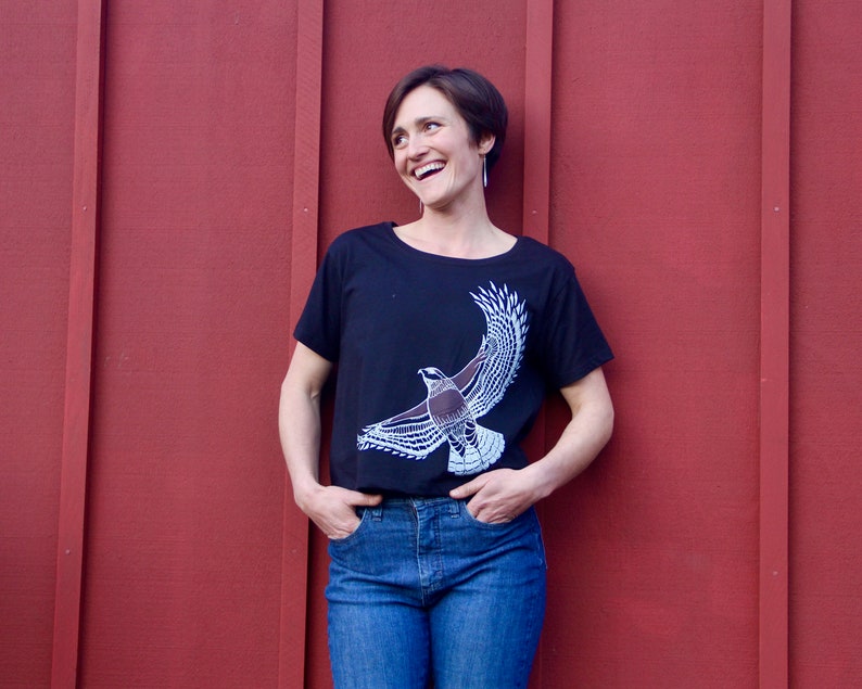 May include: A woman wearing a black t-shirt with a white bird graphic and blue jeans. She is standing in front of a red wooden wall.