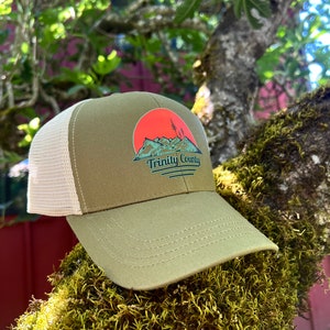 May include: A green and brown mesh trucker hat with a red sunset and mountain graphic. The text "Trinity County" is printed on the hat.