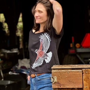 May include: A woman wearing a black t-shirt with a white and red hawk graphic. The shirt has a scoop neck and short sleeves. She is wearing blue jeans and a brown belt.