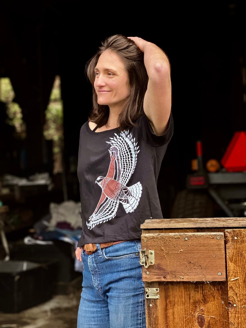 May include: A woman wearing a black t-shirt with a white and red hawk graphic. The shirt has a scoop neck and short sleeves. She is wearing blue jeans and a brown belt.
