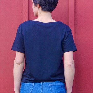 May include: A black short-sleeved t-shirt with a scoop neck. The shirt is worn by a person standing in front of a red wall.