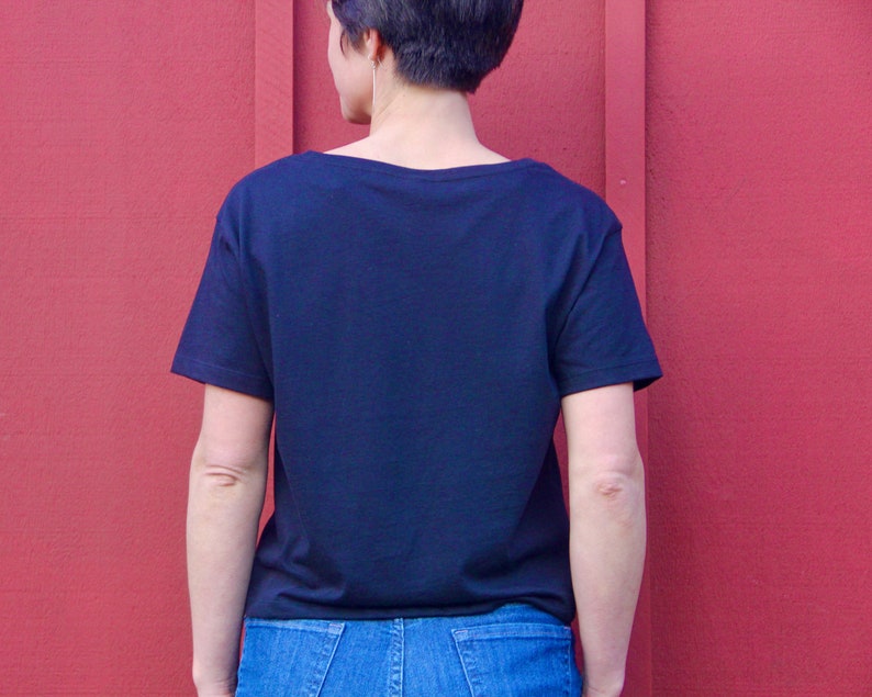 May include: A black short-sleeved t-shirt with a scoop neck. The shirt is worn by a person standing in front of a red wall.