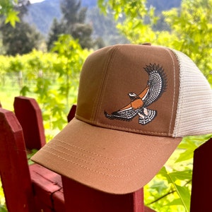 Puede incluir: Una gorra de camionero marrón y blanca con una parte delantera de lona marrón y una parte trasera de malla blanca. La gorra presenta un gráfico de un halcón en vuelo con detalles en naranja, negro y blanco.