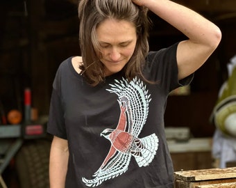 May include: A woman wearing a black cropped t-shirt with a white and red hawk design. The shirt has a relaxed fit and short sleeves. The woman is wearing blue jeans.
