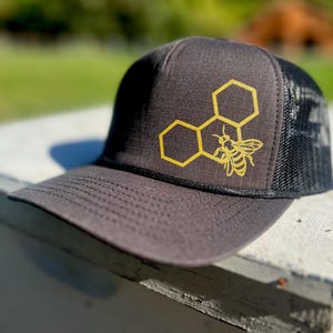 May include: A black and gray trucker hat with a gold honeycomb and bee design. The hat has a mesh back.