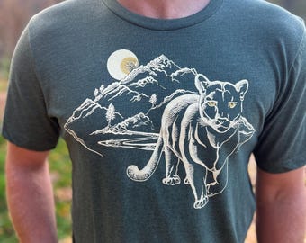Könnte beinhalten: Ein dunkelgrünes T-Shirt mit einem weißen Aufdruck einer Bergszene mit Vollmond und einem Puma, der auf den Betrachter zugeht.