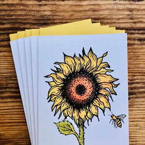 Op de afbeelding: Een set van vijf blanco wenskaarten met een handgetekende illustratie van een zonnebloem met een bij die ernaast vliegt. De zonnebloem is geel met een bruine kern en groene bladeren. De kaarten zijn wit met een gele rand.