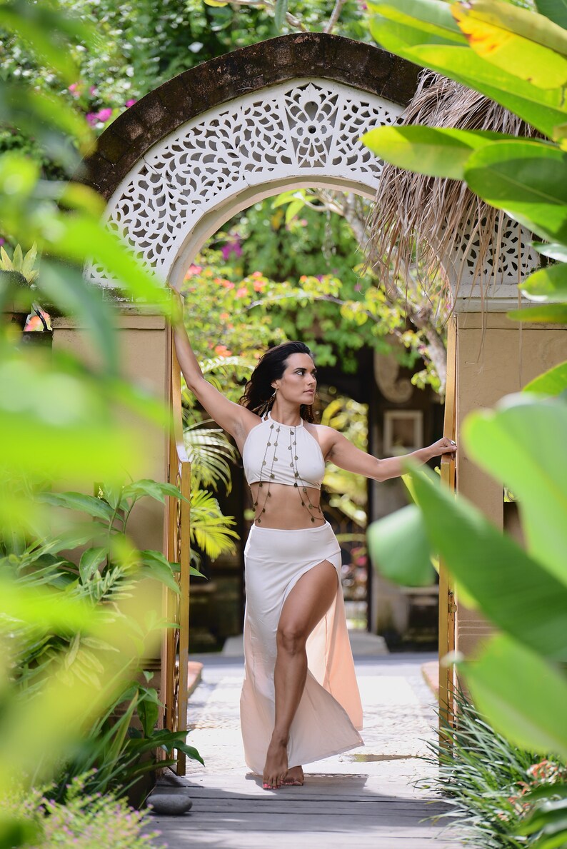 May include: A woman wearing a white halter top and a white skirt with a slit stands in a doorway with a decorative archway. The doorway is surrounded by lush green foliage.