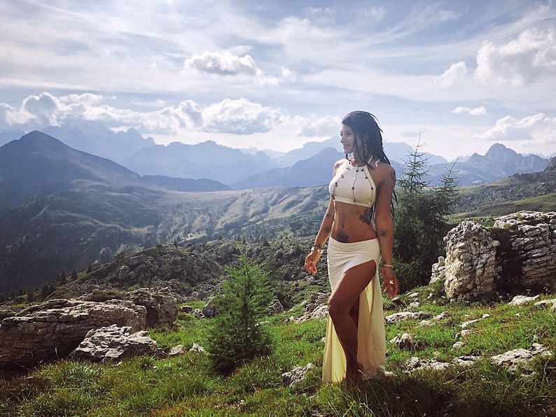 May include: A woman wearing a white halter top and a long white skirt with a slit up the side. She is standing in a mountainous landscape with a blue sky and white clouds.