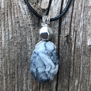 May include: A silver pendant necklace with a black cord. The pendant features a round silver bead and a teardrop-shaped grey and white stone.