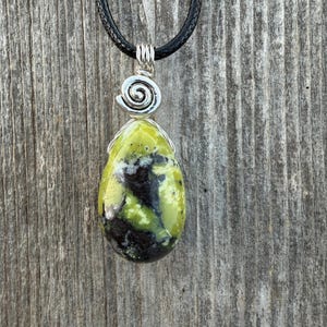 May include: A pendant necklace featuring a teardrop-shaped, green and black stone. The stone is wire-wrapped and suspended from a silver spiral design. The necklace has a black cord.