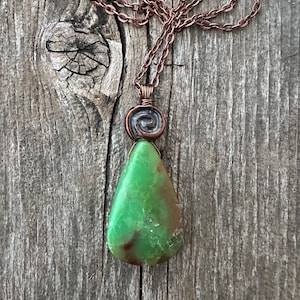 May include: A handcrafted copper necklace featuring a green gemstone pendant. The pendant is a teardrop shape with a spiral accent. The chain and wire wrapping are copper-colored, set against a weathered wood background.