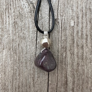 May include: A handcrafted necklace featuring a dark purple stone pendant and a silver-colored metal bead, suspended from a black leather cord. The pendant is wire-wrapped, and the necklace rests on a weathered wooden surface.