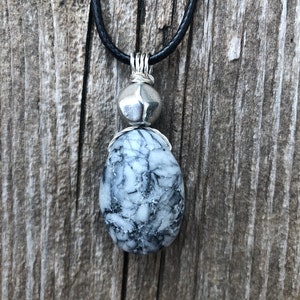 May include: A silver pendant necklace with a black cord. The pendant features a round silver bead and a larger, oval-shaped, white and grey stone with a marbled pattern.